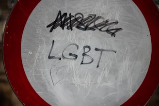 A traffic sign with a red border and a white center is defaced with graffiti. The word 'LGBT' is clearly written in black marker across the center, with additional scribbles above it.