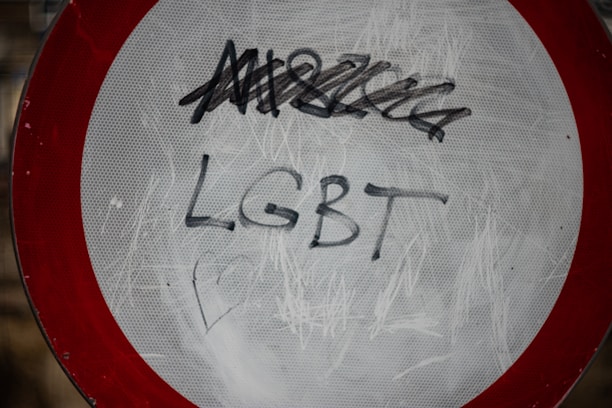 A traffic sign with a red border and a white center is defaced with graffiti. The word 'LGBT' is clearly written in black marker across the center, with additional scribbles above it.