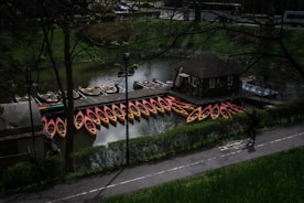 A collection of kayaks, primarily red and yellow, is lined up along a wooden dock on a calm body of water. There is a small building on the dock, likely used for rental purposes. A man is walking on a paved path near the dock, bordered by green shrubbery. The scene is shaded by tall trees with leafless branches.