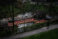A collection of kayaks, primarily red and yellow, is lined up along a wooden dock on a calm body of water. There is a small building on the dock, likely used for rental purposes. A man is walking on a paved path near the dock, bordered by green shrubbery. The scene is shaded by tall trees with leafless branches.