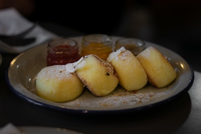 A plate with several pieces of baked goods, possibly small cakes or biscuits, lightly dusted with powdered sugar. There are three small jars of different colored jams or spreads in the background.