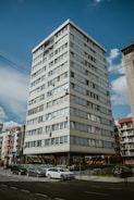 A multi-story residential building with a modern design, elevated on concrete stilts, surrounded by parked cars and adjacent to other similar structures. The sky is clear with a few scattered clouds, and there are street signs and some greenery in the vicinity.