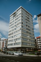 A multi-story residential building with a modern design, elevated on concrete stilts, surrounded by parked cars and adjacent to other similar structures. The sky is clear with a few scattered clouds, and there are street signs and some greenery in the vicinity.