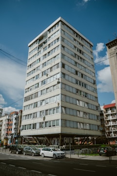 A multi-story residential building with a modern design, elevated on concrete stilts, surrounded by parked cars and adjacent to other similar structures. The sky is clear with a few scattered clouds, and there are street signs and some greenery in the vicinity.