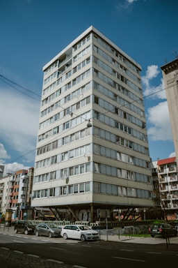 A multi-story residential building with a modern design, elevated on concrete stilts, surrounded by parked cars and adjacent to other similar structures. The sky is clear with a few scattered clouds, and there are street signs and some greenery in the vicinity.