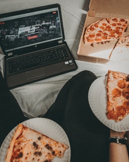 A cozy setting with a laptop displaying a streaming service interface nearby, alongside a pizza box holding a partially eaten pizza. Two slices of pizza are on paper plates held by someone, suggesting a casual movie night atmosphere.