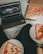 A cozy setting with a laptop displaying a streaming service interface nearby, alongside a pizza box holding a partially eaten pizza. Two slices of pizza are on paper plates held by someone, suggesting a casual movie night atmosphere.