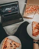 A cozy setting with a laptop displaying a streaming service interface nearby, alongside a pizza box holding a partially eaten pizza. Two slices of pizza are on paper plates held by someone, suggesting a casual movie night atmosphere.