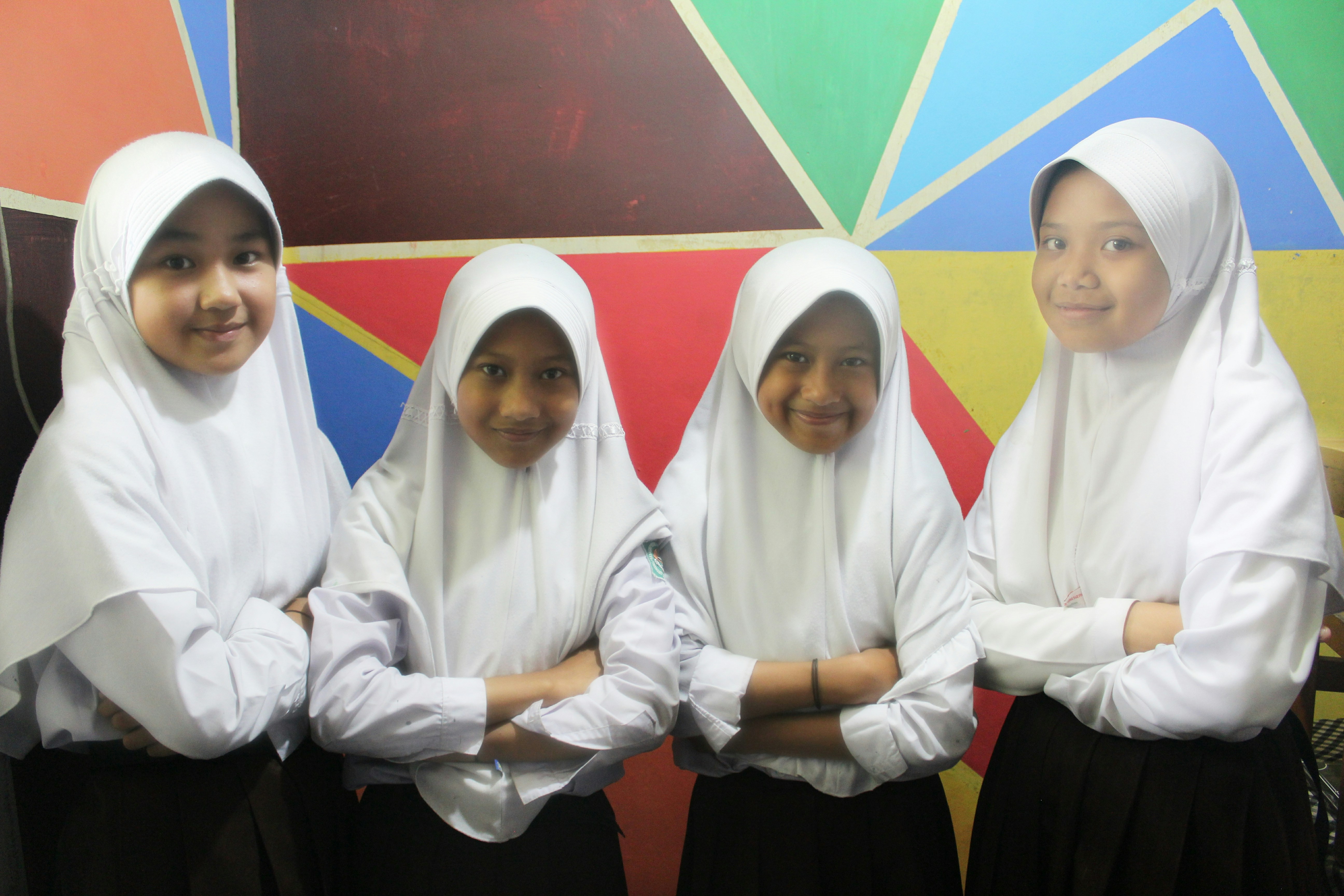 Four young girls wearing white hijabs and white blouses stand in front of a colorful geometric wall. They have their arms crossed and are smiling while facing the camera.