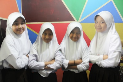 Four young girls wearing white hijabs and white blouses stand in front of a colorful geometric wall. They have their arms crossed and are smiling while facing the camera.