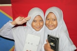 Two young girls wearing white hijabs are smiling. The girl on the left is holding a book with 'jeda' on the cover, while the girl on the right is holding a book with '00.00' on the cover. They are standing in front of a colorful backdrop featuring red, blue, and yellow sections.