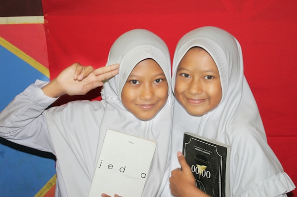 Two young girls wearing white hijabs are smiling. The girl on the left is holding a book with 'jeda' on the cover, while the girl on the right is holding a book with '00.00' on the cover. They are standing in front of a colorful backdrop featuring red, blue, and yellow sections.