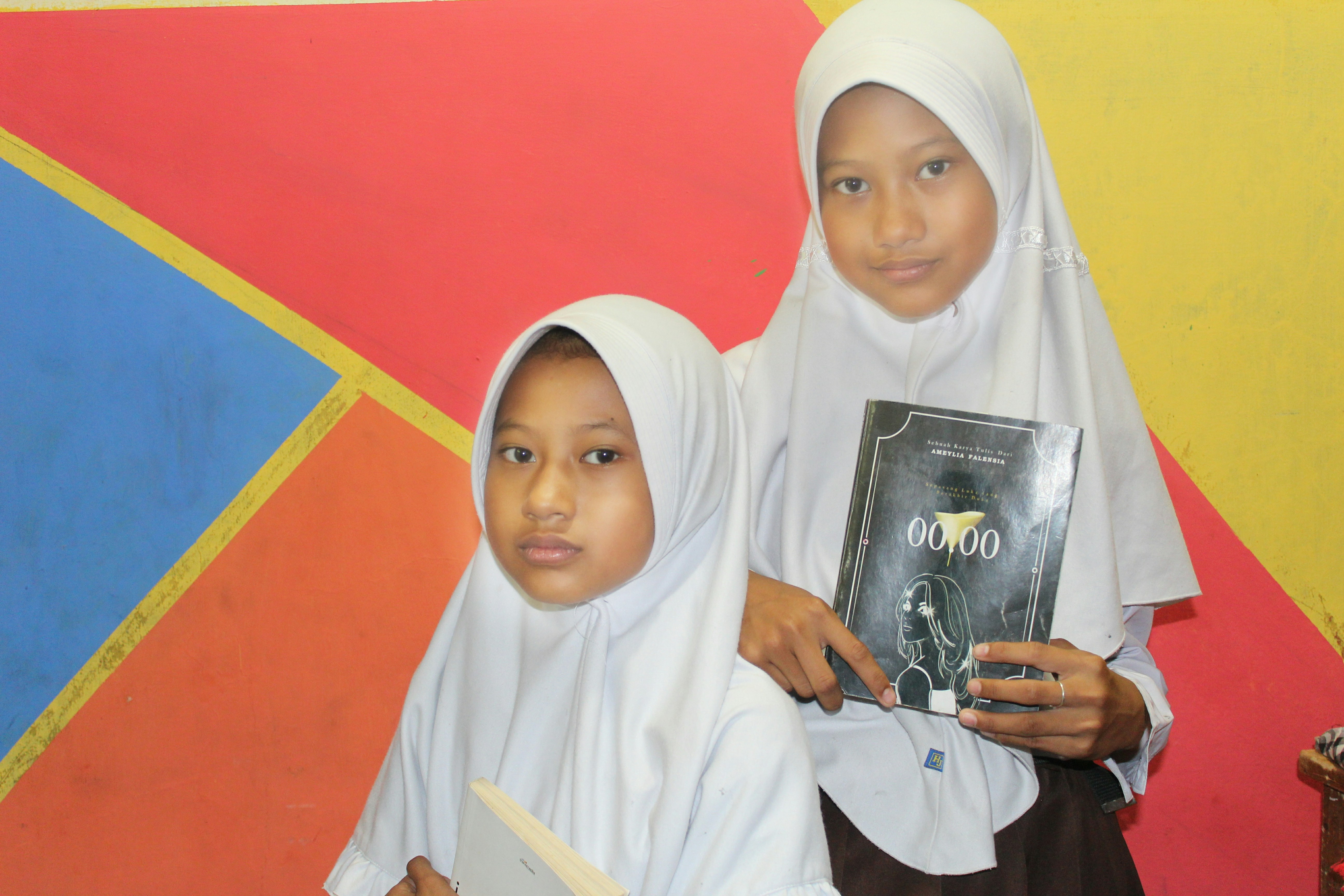Two young girls wearing white hijabs hold books while standing in front of a colorful geometric background featuring red, blue, orange, and yellow sections.