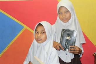 Two young girls wearing white hijabs hold books while standing in front of a colorful geometric background featuring red, blue, orange, and yellow sections.