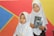 Two young girls wearing white hijabs hold books while standing in front of a colorful geometric background featuring red, blue, orange, and yellow sections.