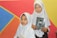 Two young girls wearing white hijabs hold books while standing in front of a colorful geometric background featuring red, blue, orange, and yellow sections.