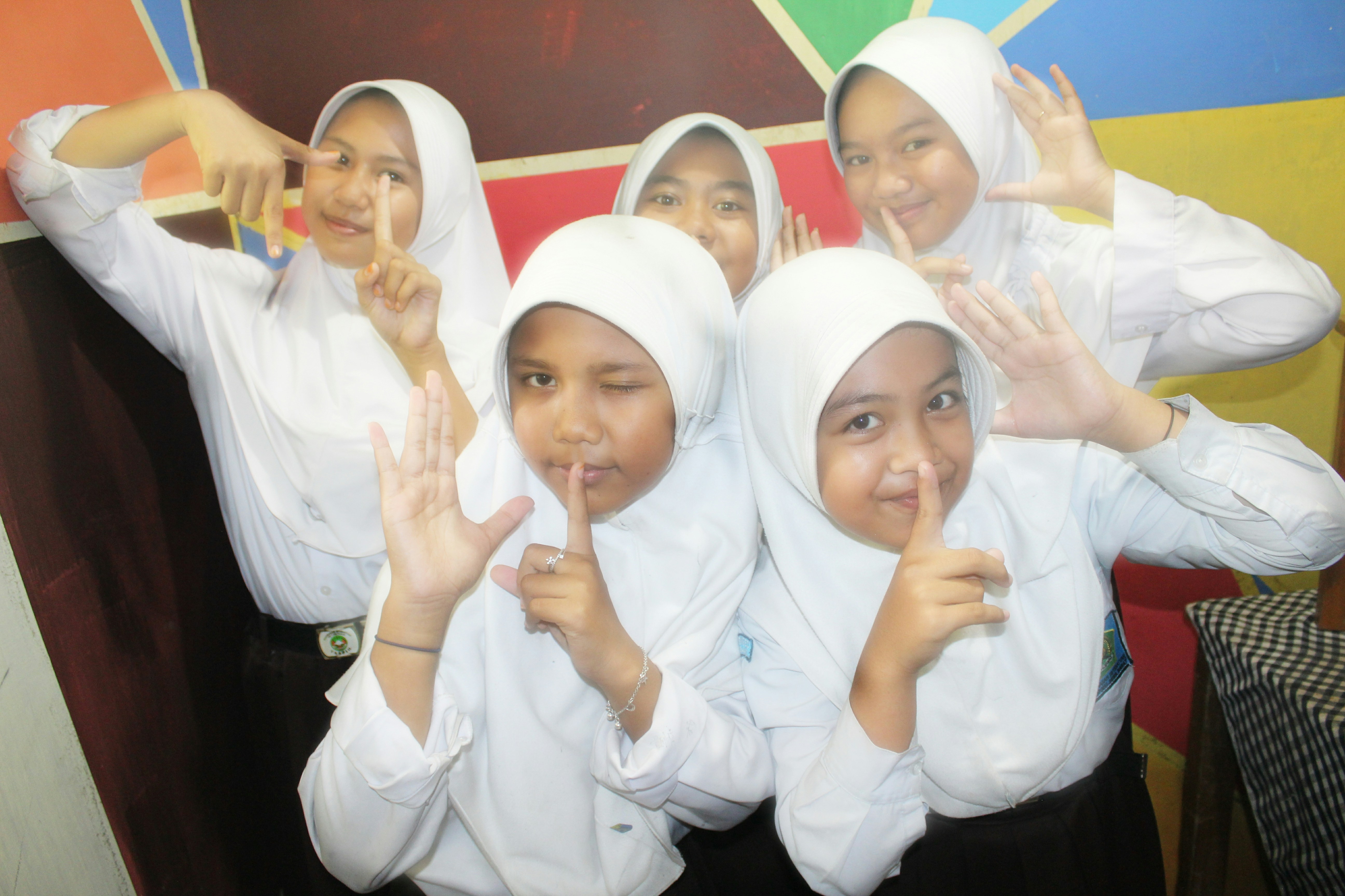 Five young individuals wearing white uniforms and hijabs are posing playfully, with their hands forming gesture signs in front of a colorful geometric wall.