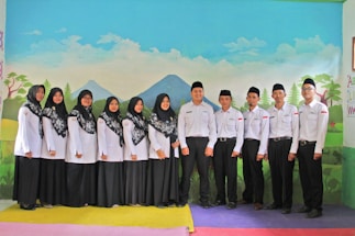 A group of men and women dressed in matching uniforms, consisting of white tops and black skirts or pants, are standing indoors in front of a painted backdrop depicting a landscape with mountains, trees, and clouds. The men wear black hats, and the women are wearing headscarves with intricate patterns.