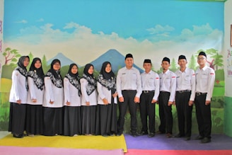 A group of men and women dressed in matching uniforms, consisting of white tops and black skirts or pants, are standing indoors in front of a painted backdrop depicting a landscape with mountains, trees, and clouds. The men wear black hats, and the women are wearing headscarves with intricate patterns.