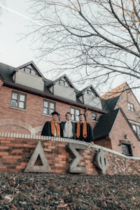 Three graduates in academic gowns and caps stand in front of a brick building featuring Greek letters on the wall. The setting appears to be a college campus, and the trees surrounding the area are leafless, suggesting fall or winter.