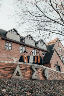 Three graduates in academic gowns and caps stand in front of a brick building featuring Greek letters on the wall. The setting appears to be a college campus, and the trees surrounding the area are leafless, suggesting fall or winter.