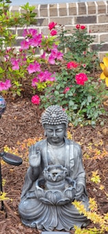 A serene garden scene featuring a Buddha statue seated in a meditative pose, surrounded by vibrant pink and red flowers. The statue is set on a bed of brown mulch, with a small light fixture nearby, and brick steps in the background.