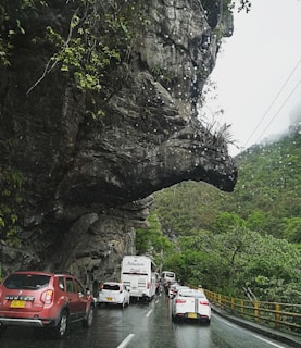A road winds through a lush, mountainous region with a distinctive rock formation resembling an animal's head protruding over the road. Cars and buses are seen traveling along the wet roadway, suggesting recent rain. The scenery is rich with greenery, including trees and foliage, and the sky is overcast with light rain.