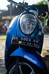A blue scooter with a detailed focus on the front, including its headlight and a visible license plate marked DK 5003 VW. The scooter also features a sticker that reads 'gotrovela'. Sunlight enhances the scene, casting shadows and highlighting the metallic blue color of the scooter. The background is slightly blurred, suggesting an outdoor, possibly urban, setting.