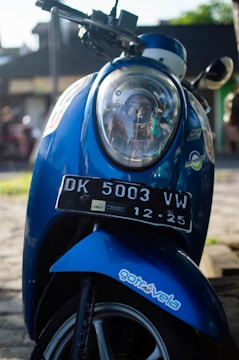 A blue scooter with a detailed focus on the front, including its headlight and a visible license plate marked DK 5003 VW. The scooter also features a sticker that reads 'gotrovela'. Sunlight enhances the scene, casting shadows and highlighting the metallic blue color of the scooter. The background is slightly blurred, suggesting an outdoor, possibly urban, setting.