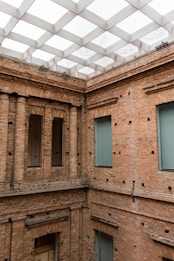 An interior courtyard with exposed brick walls and rectangular windows. The ceiling features a modern, grid-like geometric structure with numerous recessed transparent panels that provide natural light. The architectural design combines classical and contemporary elements with a focus on the brickwork and open space.