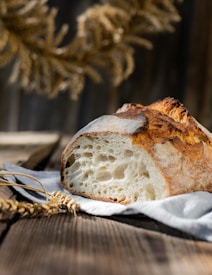 A rustic loaf of bread with a crusty, golden-brown exterior sits on a wooden table. The bread is sliced to show a soft, airy interior, revealing large bubbles indicative of an artisanal baking style. In the foreground, there are wheat stalks, and a sunlit, blurred background gives a warm, natural feel.