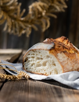 A rustic loaf of bread with a crusty, golden-brown exterior sits on a wooden table. The bread is sliced to show a soft, airy interior, revealing large bubbles indicative of an artisanal baking style. In the foreground, there are wheat stalks, and a sunlit, blurred background gives a warm, natural feel.
