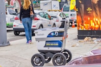 A delivery robot is on a city sidewalk alongside a woman walking away. The background shows a variety of vehicles, including a white car and a U-Haul van, with some street clutter and a large advertisement poster featuring candles.