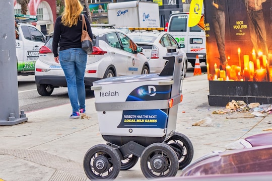 A delivery robot is on a city sidewalk alongside a woman walking away. The background shows a variety of vehicles, including a white car and a U-Haul van, with some street clutter and a large advertisement poster featuring candles.