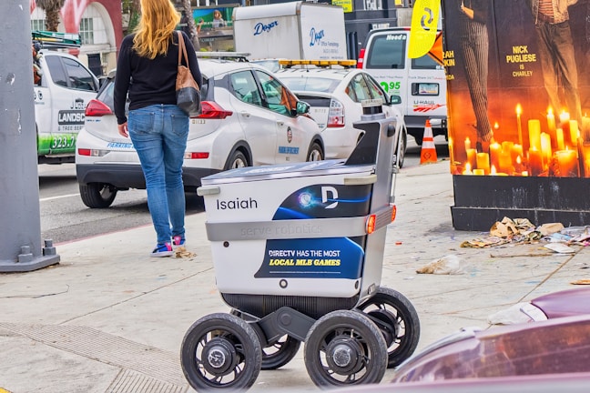 A delivery robot is on a city sidewalk alongside a woman walking away. The background shows a variety of vehicles, including a white car and a U-Haul van, with some street clutter and a large advertisement poster featuring candles.