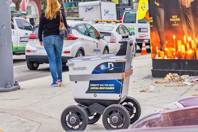 A delivery robot is on a city sidewalk alongside a woman walking away. The background shows a variety of vehicles, including a white car and a U-Haul van, with some street clutter and a large advertisement poster featuring candles.