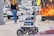 A delivery robot is on a city sidewalk alongside a woman walking away. The background shows a variety of vehicles, including a white car and a U-Haul van, with some street clutter and a large advertisement poster featuring candles.