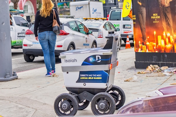 A delivery robot is on a city sidewalk alongside a woman walking away. The background shows a variety of vehicles, including a white car and a U-Haul van, with some street clutter and a large advertisement poster featuring candles.