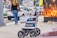 A delivery robot is on a city sidewalk alongside a woman walking away. The background shows a variety of vehicles, including a white car and a U-Haul van, with some street clutter and a large advertisement poster featuring candles.