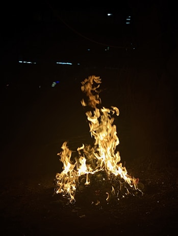 A vivid blaze of flames flickers brightly in a dark setting, casting an intense glow from the burning pile of material. The fire twists and turns, showing dynamic movement and energy.