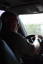 A person is driving a vehicle, holding the steering wheel with both hands. The focus is on the driver who is wearing glasses and a wristwatch, with light streaming in from a window. There are green trees visible outside through the windshield.