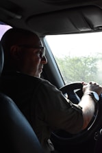 A person is driving a vehicle, holding the steering wheel with both hands. The focus is on the driver who is wearing glasses and a wristwatch, with light streaming in from a window. There are green trees visible outside through the windshield.