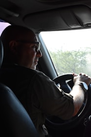 A person is driving a vehicle, holding the steering wheel with both hands. The focus is on the driver who is wearing glasses and a wristwatch, with light streaming in from a window. There are green trees visible outside through the windshield.