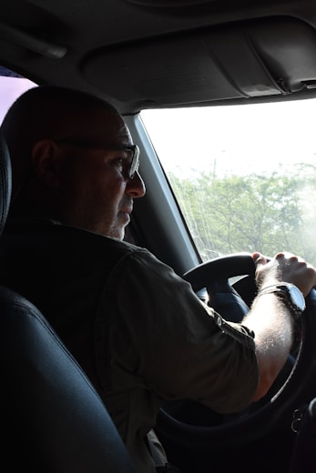 A person is driving a vehicle, holding the steering wheel with both hands. The focus is on the driver who is wearing glasses and a wristwatch, with light streaming in from a window. There are green trees visible outside through the windshield.