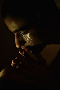 A contemplative individual in low light, with a soft spotlight highlighting their face. Their eyes are closed, and there are visible tear tracks on their cheek, suggesting deep emotion. Their hands are clasped near their mouth, contributing to a sense of introspection.