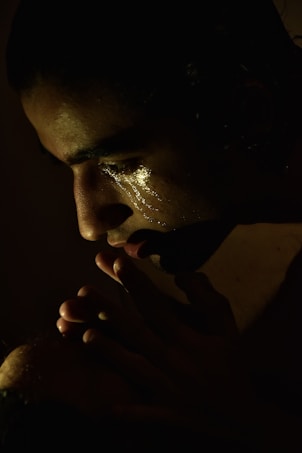 A contemplative individual in low light, with a soft spotlight highlighting their face. Their eyes are closed, and there are visible tear tracks on their cheek, suggesting deep emotion. Their hands are clasped near their mouth, contributing to a sense of introspection.