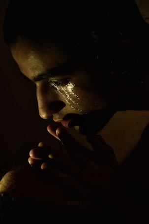 A contemplative individual in low light, with a soft spotlight highlighting their face. Their eyes are closed, and there are visible tear tracks on their cheek, suggesting deep emotion. Their hands are clasped near their mouth, contributing to a sense of introspection.