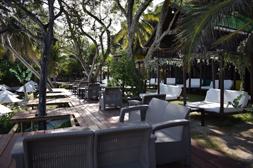 A scenic outdoor lounge area featuring wicker furniture set on a wooden deck amidst lush greenery. White lounge chairs and sofas are arranged neatly, offering a comfortable seating space under the shade of tall palm trees. Decorative hanging plants add to the tropical ambiance.