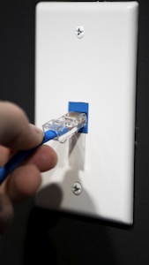 A hand is inserting a blue Ethernet cable into a white wall socket. The socket and cable appear to be part of a network setup.