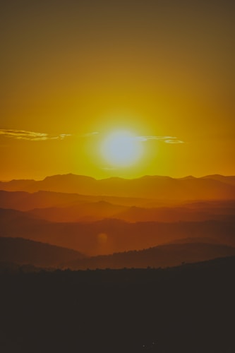 The image captures a warm and golden sunset over a series of rolling hills and mountains. The sun casts a vibrant glow, creating a layered silhouette effect with the landscape. Wisps of clouds are visible in the sky, adding texture to the scene.